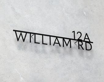 Pode incluir: Placa de endereço de metal preto com o texto "WILLIAM RD 12A" em uma fonte moderna. A placa é montada em uma parede texturizada cinza claro, criando uma estética minimalista.