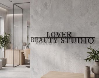 May include: A black metal wall sign featuring the text LOVER BEAUTY STUDIO in a serif font. The sign is mounted horizontally on a light gray textured concrete wall, creating a clean and professional display for a business interior space.
