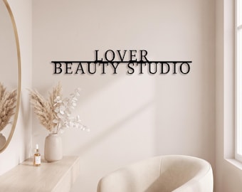 May include: A black metal wall sign featuring the text LOVER BEAUTY STUDIO in a clean, sans-serif font. The letters are connected by a horizontal line that runs beneath the word LOVER and above the word BEAUTY STUDIO, creating a unified piece.