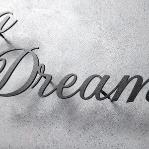 May include: Black metal wall art spelling out the word "Dream" in a cursive font. The letters are mounted on a textured, light gray surface, creating a contrast between the dark text and the background.