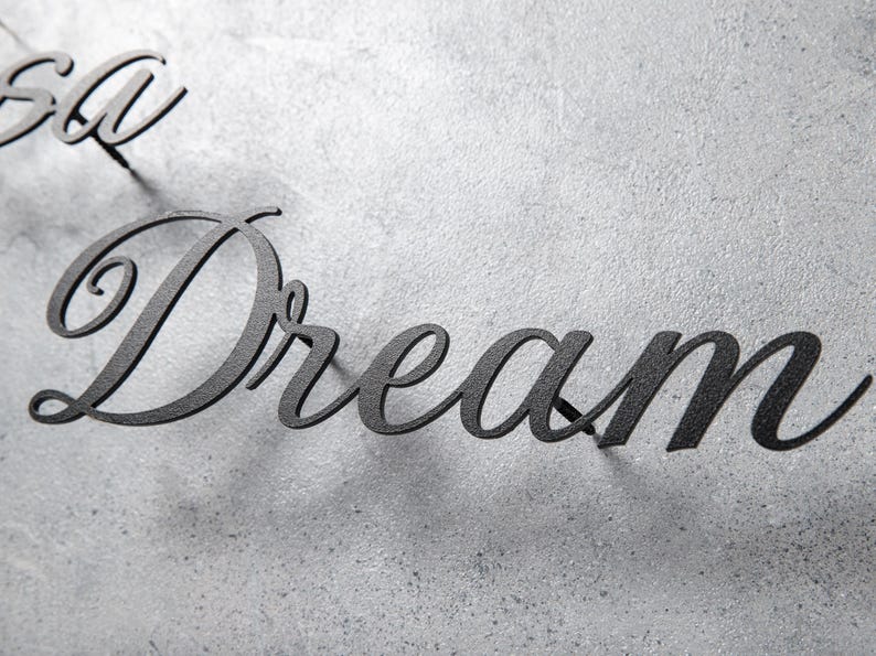 May include: Black metal wall art spelling out the word "Dream" in a cursive font. The letters are mounted on a textured, light gray surface, creating a contrast between the dark text and the background.