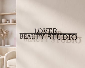 May include: A black metal wall sign featuring the text LOVER BEAUTY STUDIO in a bold, sans-serif font. The sign is mounted on a light-colored wall, creating a distinct shadow behind the letters. The design consists of two lines of text.