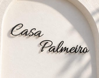 May include: Black metal wall lettering spelling out the words Casa Palmeiro in a cursive script font. The letters are mounted on a textured off-white wall surface that features a large, recessed archway design surrounding the text.