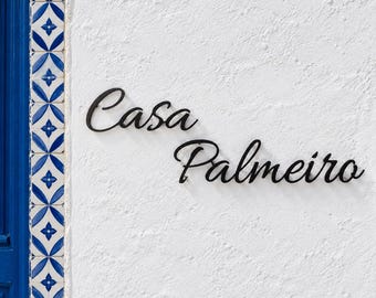 May include: A black metal wall sign featuring the cursive text Casa Palmeiro mounted on a white textured wall next to a vertical border of blue and white patterned tiles. The sign is composed of two separate words in a script font.