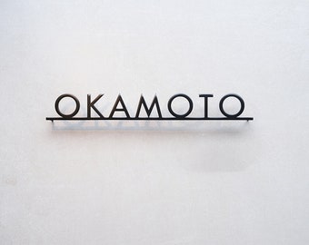 Pode incluir: Uma placa de parede em metal preto com a palavra OKAMOTO em letras maiúsculas. As letras estão ligadas por uma fina barra horizontal ao longo da base, criando um design linear e limpo que se destaca na parede de cor clara.
