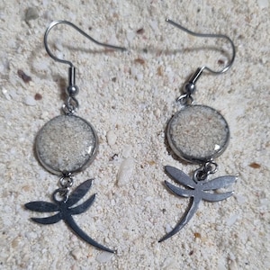 May include: Silver-tone drop earrings with a circular pendant filled with sand and a dragonfly charm. The earrings have a hook fastening. The earrings are displayed on a sandy surface. The earrings are a beach-themed accessory.