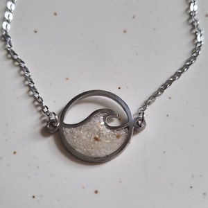 Könnte beinhalten: Ein silbernes Kettenarmband mit einem runden Anhänger, der ein Wellenmuster zeigt, das mit weißem Sand gefüllt ist.
