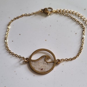 Könnte beinhalten: Ein goldenes Kettenarmband mit einem runden Anhänger, der ein Wellenmuster zeigt, das mit weißem Sand gefüllt ist.