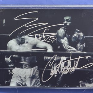 Puede incluir: Fotografía en blanco y negro de dos boxeadores en un ring de boxeo. Ambos boxeadores están sin camisa y llevan pantalones cortos de boxeo. La imagen está firmada con dos autógrafos en tinta blanca.