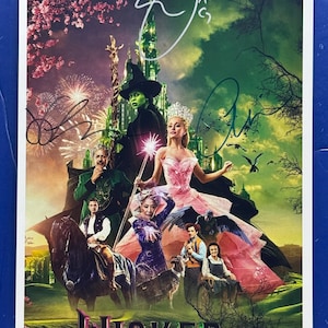 Puede incluir: Un póster del musical "Wicked" con una foto del elenco y firmas. La imagen muestra personajes disfrazados contra un telón de fondo de un castillo verde, fuegos artificiales y un cielo colorido. La palabra "Wicked" está en la parte inferior.
