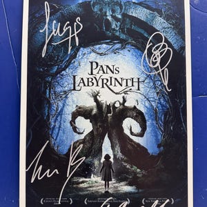 May include: A movie poster for Pan's Labyrinth featuring a dark, blue-toned forest scene with a central stone archway and a small figure standing before it. The poster includes the title, text in German, and several handwritten signatures in white ink.