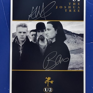 以下が含まれることがあります： バンドU2のモノクロ写真が印刷された長方形のポスター。金色の文字で「U2 THE JOSHUA TREE」と記されている。画像には銀色のインクで書かれた2つのサインが見える。下部には金色の木のアートとU2のロゴが配置されている。