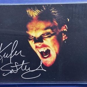 Könnte beinhalten: Signiertes Foto mit einem Nahaufnahme von Kiefer Sutherland mit bedrohlichem Gesichtsausdruck, gebleckten Zähnen und leuchtenden Augen. Die Signatur "Kiefer Sutherland" ist in weißer Tinte sichtbar. Das Bild hat einen dunklen Hintergrund mit rotem und gelbem Farbschema.