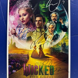 Puede incluir: Un póster promocional del musical "Wicked" con varias firmas. El póster presenta un elenco de personajes y el texto "Wicked For Good" y "November 21". El fondo presenta un paisaje de fantasía con un camino de ladrillos amarillos.