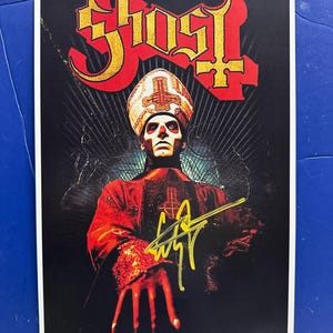 May include: A poster featuring the band Ghost. The poster has the band's logo in gold and red at the top. The image shows a person in a red and black robe and a white hat with a cross. The image is signed in yellow.