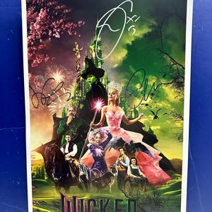May include: A vertical movie poster for Wicked featuring a central green-skinned witch and a blonde witch in a pink gown, surrounded by supporting characters, a horse, and a castle. The poster includes three distinct black ink signatures.