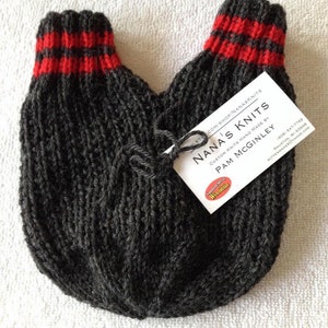May include: A pair of grey knitted mittens with red and black stripes at the cuffs. The mittens are handmade by Nana's Knits, a custom knitter in Mauston, Wisconsin.