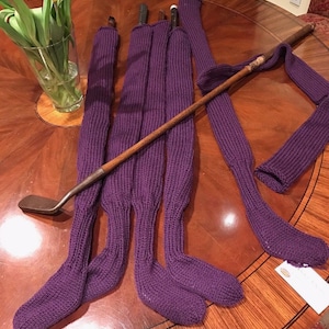 May include: Five purple knitted golf club covers with a wooden golf club lying on top of them. The golf club covers are on a wooden table.