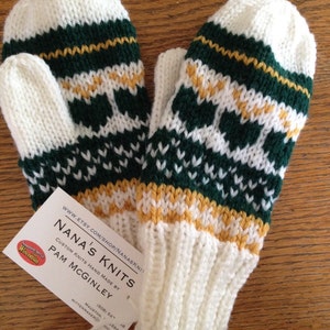 May include: A pair of white knit mittens with green, white, and gold stripes. The mittens are handmade and have a tag that says "Nana's Knits" and "Custom Knits Hand Made By Pam McGinley".