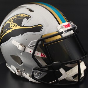 May include: Silver football helmet with a black facemask and visor. The helmet features a gold and teal stripe and a black and gold jaguar logo on the side. The Riddell logo is visible on the visor and chin strap.