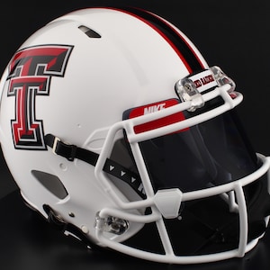 Puede incluir: Un casco de fútbol americano blanco con una franja negra y roja y el logotipo de los Red Raiders de Texas Tech. El casco tiene una visera negra, una máscara facial blanca y el logotipo de Nike. Las palabras "Red Raiders" son visibles en el clip de la visera.