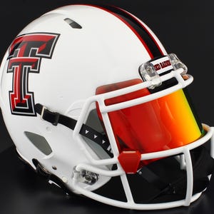 Puede incluir: Casco de fútbol americano blanco con una raya roja y negra y una visera roja. El casco presenta un gran logotipo "T" rojo con un contorno negro. La máscara facial es blanca y la visera está teñida de rojo y amarillo.