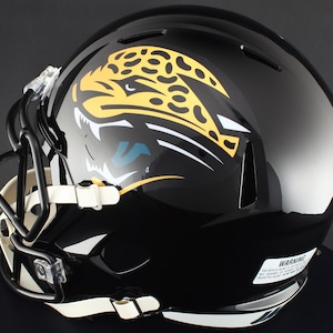 May include: A glossy black football helmet featuring a large yellow and black jaguar head logo on the side. The helmet includes a black face mask, white chin straps, and a white warning label on the lower back edge with visible black text.
