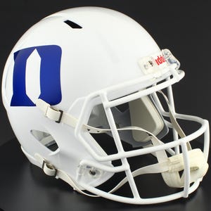 May include: A white football helmet with a blue "D" logo on the side. The helmet has a white face mask, chin strap, and clear plastic clips. The helmet is designed for protection during American football games.