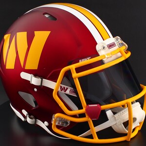 May include: A burgundy American football helmet with a yellow and white stripe design. The helmet features a yellow "W" logo and a yellow facemask. The visor is tinted, and the helmet has the word "Commanders" on the visor clip.