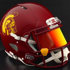 Könnte beinhalten: Ein weinroter American-Football-Helm mit einem gelb-weinroten Trojaner-Logo. Der Helm hat ein weinrotes Gesichtsgitter und ein gelb getöntes Visier. Der Helm steht auf einer schwarzen Oberfläche.