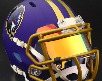 May include: A purple football helmet with gold stripes and a gold facemask. The helmet features a team logo on the side and a tinted visor. The chin strap is white. The helmet is designed for protection during American football games.