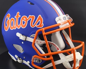 Op de afbeelding: Een blauwe American footballhelm met oranje en witte accenten. Het woord "Gators" staat in het oranje geschreven. De helm heeft een oranje gezichtsmasker, een witte kinband en een witte streep in het midden.