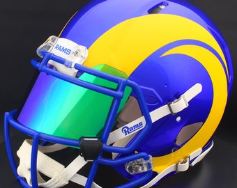 Puede incluir: Casco de fútbol americano azul y amarillo con visera verde. El casco tiene la palabra "Rams" en el lateral y en la parte delantera. Acabado brillante y correas blancas.