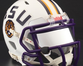 Puede incluir: Casco de fútbol americano blanco con máscara facial y visera moradas. El casco presenta las letras "SU" y un logotipo de tigre. Rayas naranjas y moradas recorren la parte superior. El logotipo de Riddell es visible en la visera.