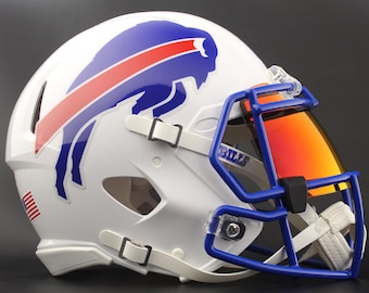 Puede incluir: Casco de fútbol americano blanco con un logotipo de búfalo azul y rojo. El casco tiene una máscara azul, una visera roja y la palabra "BILLS" es visible. Incluye una correa para la barbilla y está diseñado para la protección durante el juego.