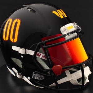 May include: A black American football helmet featuring the number "00" in orange and yellow. The helmet has a red and yellow visor, a black facemask, and the Commanders logo. The helmet is glossy and appears to be new.