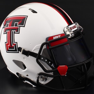 Puede incluir: Casco de fútbol americano blanco con una máscara negra y un logotipo rojo y negro. El casco tiene una raya roja en el centro y la palabra "NIKE" es visible. La visera está tintada y el casco tiene una correa para la barbilla.