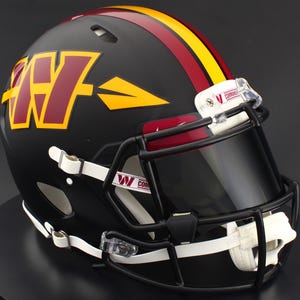 May include: A matte black football helmet featuring a burgundy and yellow W logo with an arrow design on the side. The helmet has a burgundy and yellow center stripe, a black face mask, a dark tinted visor, and white chin straps with Commanders text.