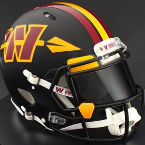 May include: A matte black football helmet featuring a burgundy and gold W logo on the side with an arrow design. The helmet includes a black face mask, a dark tinted visor, and white chin straps. The word COMMANDERS is printed on the visor clips.