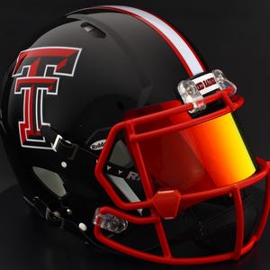 Puede incluir: Un casco de fútbol americano negro con una franja roja y blanca y una máscara facial roja. El casco presenta un gran logotipo "T" rojo con ribete blanco y una visera. La visera es un degradado de amarillo a naranja. El texto "RED RAIDERS" es visible.