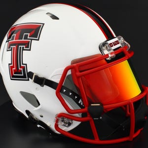 Puede incluir: Casco de fútbol americano blanco con un logotipo rojo y negro. Cuenta con una máscara facial roja y una visera tintada. Diseñado para la protección durante los partidos de fútbol americano.