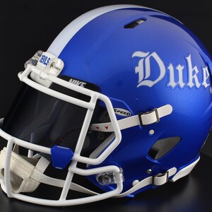 May include: A glossy blue football helmet featuring a white center stripe and the word Duke in white lettering on the side. The helmet includes a white face mask, a dark tinted visor with the word Nike, and a chin strap with the word Devils.