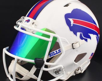 Puede incluir: Casco de fútbol americano blanco con un diseño de rayas azules y rojas. Presenta un logotipo de búfalo azul y la palabra "BILLS" en azul. La visera es verde y el casco tiene una máscara facial y una correa para la barbilla blancas.