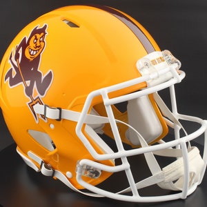 May include: A gold and brown football helmet with a white face mask. The helmet features a maroon stripe and a cartoon devil logo on the side. The helmet is designed for protection during American football games.