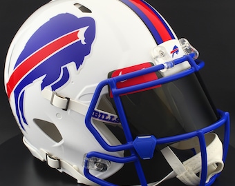 Puede incluir: Casco de fútbol americano blanco con rayas azules y rojas. Presenta un logotipo de búfalo azul y una máscara facial azul. La palabra "BILLS" es visible en la máscara facial. El casco tiene una visera tintada y una correa de barbilla blanca.