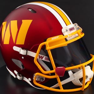 May include: A burgundy American football helmet with a yellow and white stripe and a yellow facemask. The helmet features a large yellow "W" logo and the word "Commanders" on the visor.