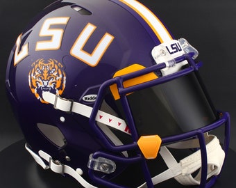 May include: A purple American football helmet with white and orange detailing. The helmet displays the letters "LSU", a tiger logo, and a white stripe. The face mask is purple with a black visor.
