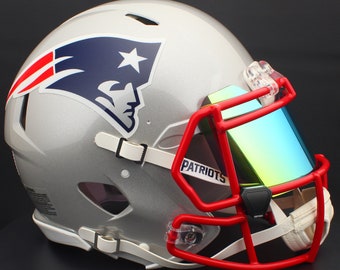 Puede incluir: Casco de fútbol americano plateado con el logotipo de los New England Patriots. El casco tiene una máscara roja, una visera con tinte arcoíris y la palabra "PATRIOTS" en la máscara.