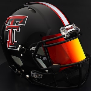 Puede incluir: Casco de fútbol americano negro con una franja roja y blanca y una visera roja. El casco presenta un gran logotipo "T" rojo y blanco y las palabras "Red Raiders" en el clip de la visera.