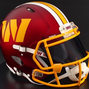 May include: A burgundy American football helmet featuring a yellow 'W' logo and a yellow stripe. The helmet has a yellow faceguard and a dark visor. The word 'Commanders' is visible on the visor clip.